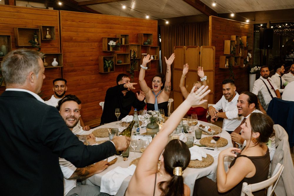 Guests cheer and raise their arms at a long dining table with lit candles and glasses during a lively wedding reception dinner.