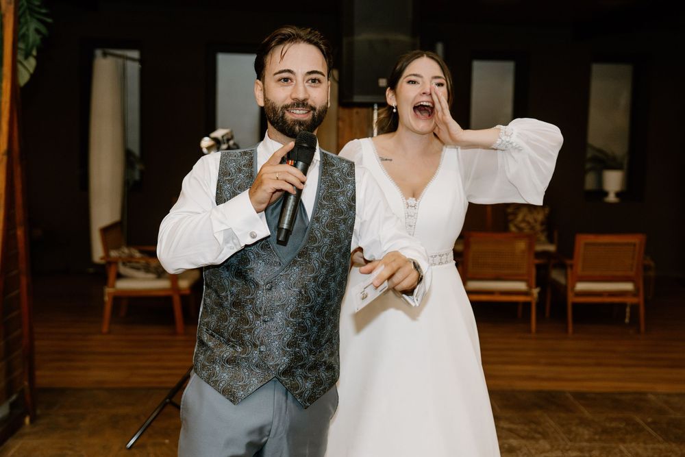 Smiling groom in a gray vest and white shirt holds a microphone beside a laughing bride in a white gown during a wedding reception toast.