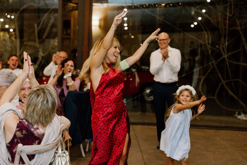 Woman in a red dress dances with raised arms at a festive indoor party while a little girl in white dances nearby and guests clap.