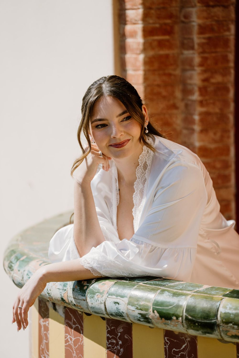 Smiling woman in a white lace-trimmed robe leans on a green tiled balcony ledge with a brick wall behind her.