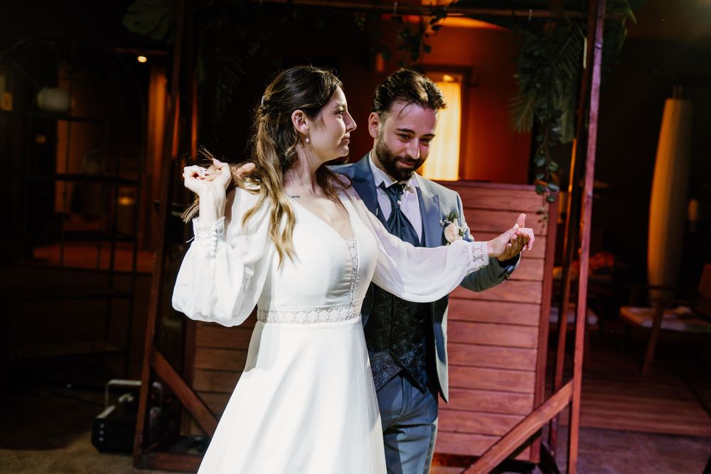 A bride in a white long-sleeved gown and a bearded groom in a blue suit share a joyful dance at their wedding reception.