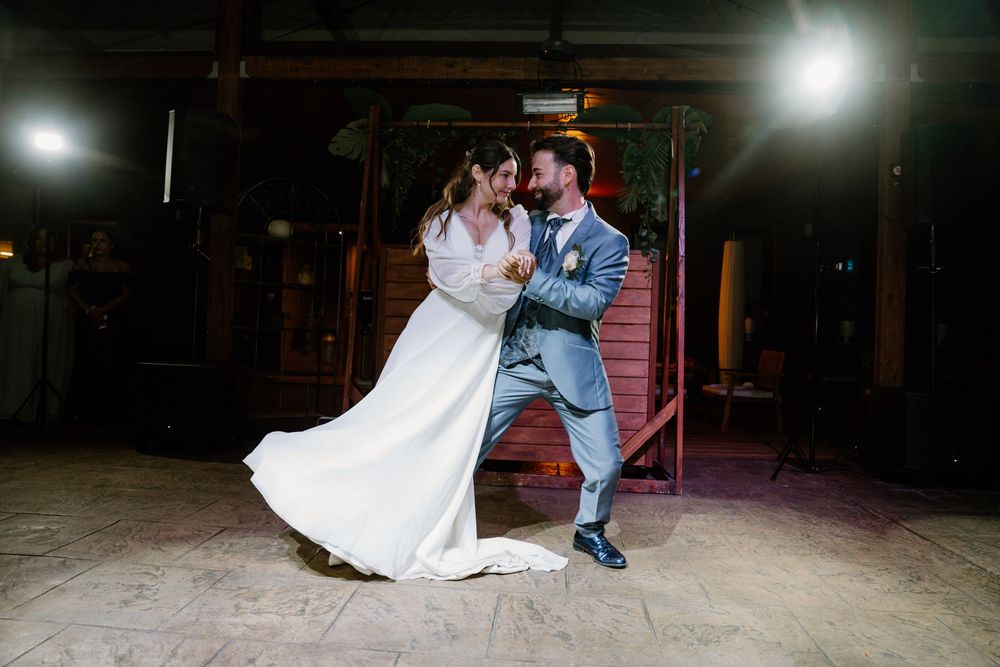 Bride in a white gown and groom in a blue suit share a romantic first dance on a rustic wooden floor with dramatic spotlights.