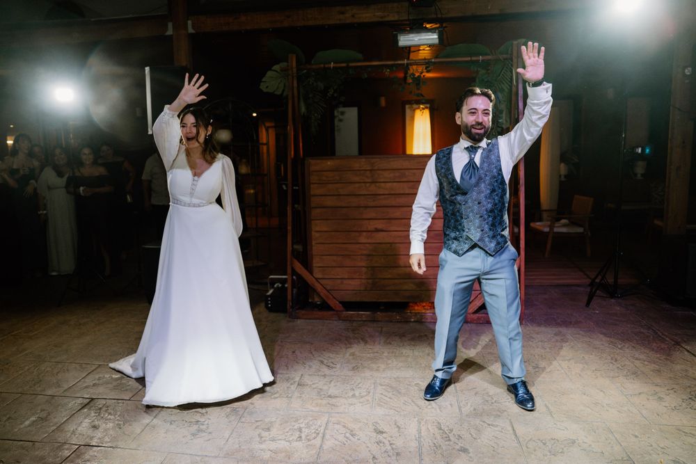 A bride in a flowing white gown and a groom in a vest and tie raise their hands during a wedding reception dance on a rustic wooden floor.