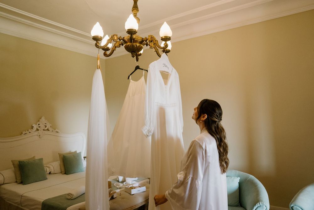Bride in a white robe looking at her wedding dress and veil hanging from a chandelier in a elegant bedroom.