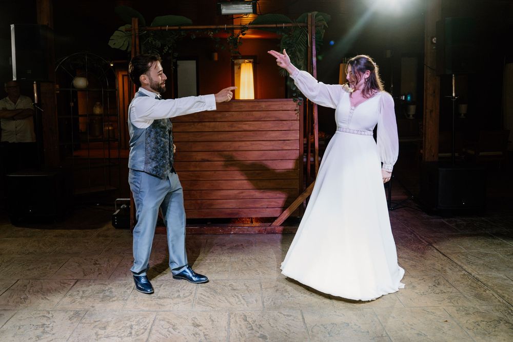 Bride in a white gown and groom in a vest and gray trousers share their first dance on a rustic wooden venue floor.
