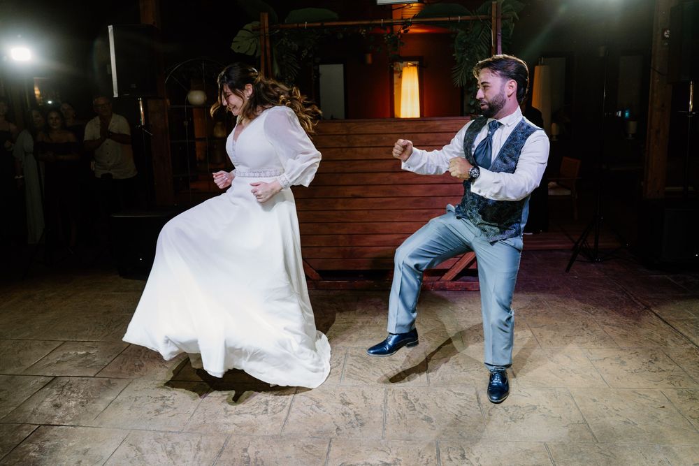 A bride in a flowing white gown and a groom in a vest and blue trousers dance energetically at their rustic wedding reception.