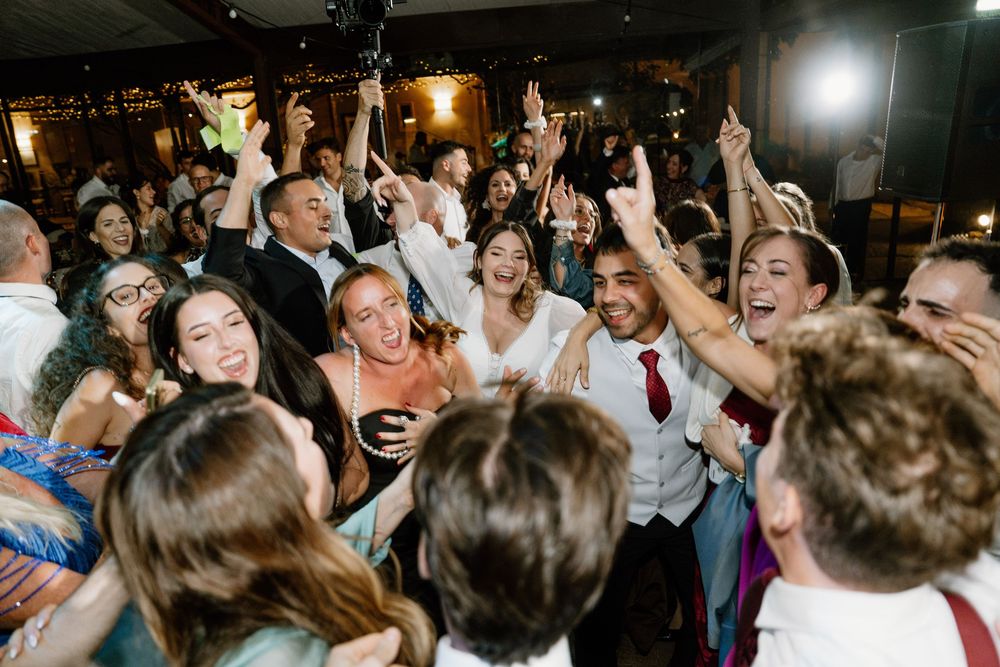 Joyful wedding guests dance with raised hands in a crowded, warmly lit reception hall, celebrating together on the dance floor.
