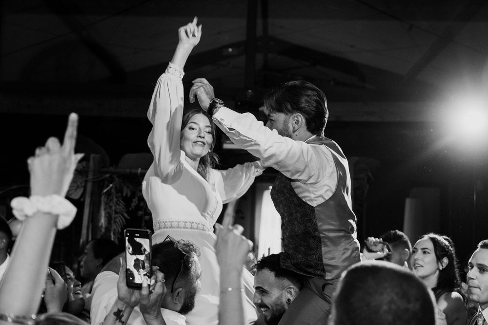 A bride in a white dress raises her arm and dances joyfully with a man in a vest as guests cheer at an evening wedding party.