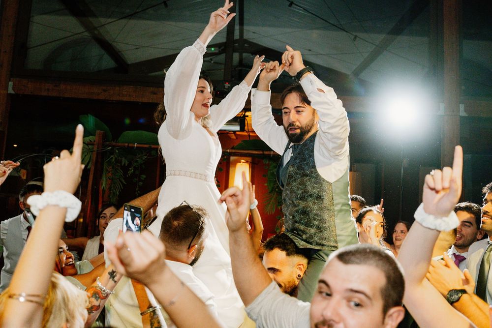 Wedding guests lift a bride in a white dress and a groom in a green vest on their shoulders as the crowd cheers and claps.