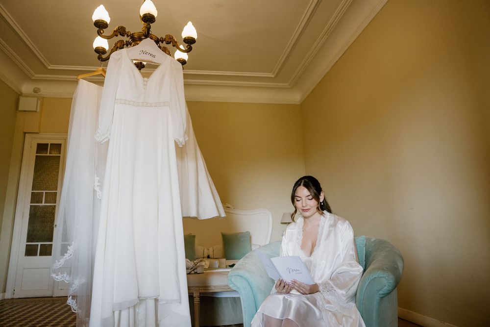 Bride in a white satin robe sits in a teal armchair reading a letter, with her wedding dress hanging from a chandelier in an elegant room.