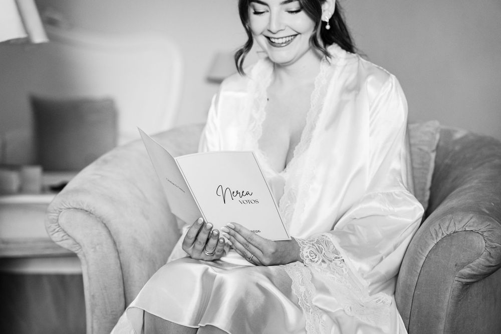 Smiling bride in a white satin robe sits on a sofa reading a card in black and white.