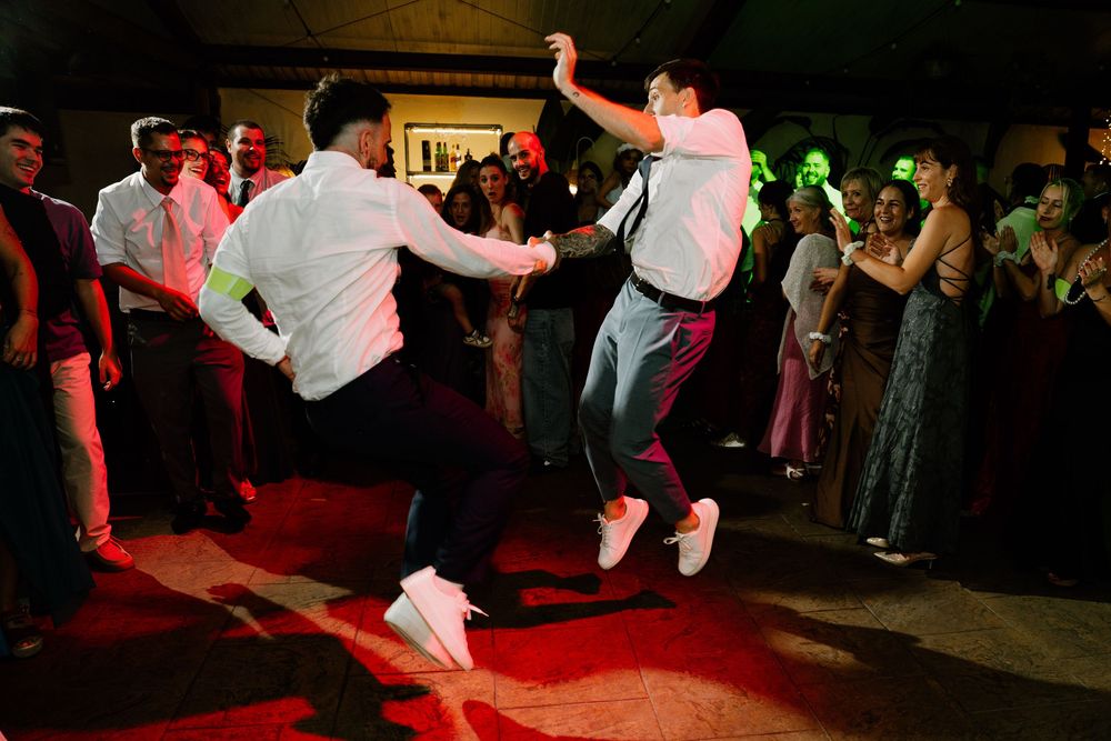 Two wedding guests dance energetically on a red-lit dance floor while a cheering crowd of guests watches and claps behind them.