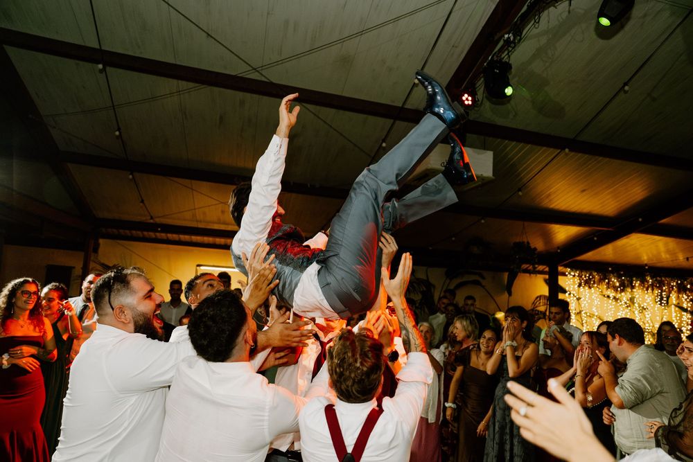 Wedding guests lift a man into the air above the crowd in a rustic, beam-ceilinged reception hall during a lively celebration.