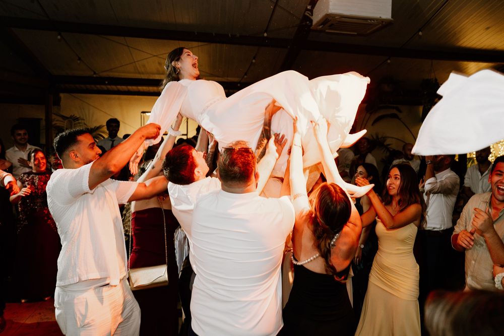 Wedding guests lift a person in a white dress into the air as the crowd cheers and celebrates on a lively reception dance floor.
