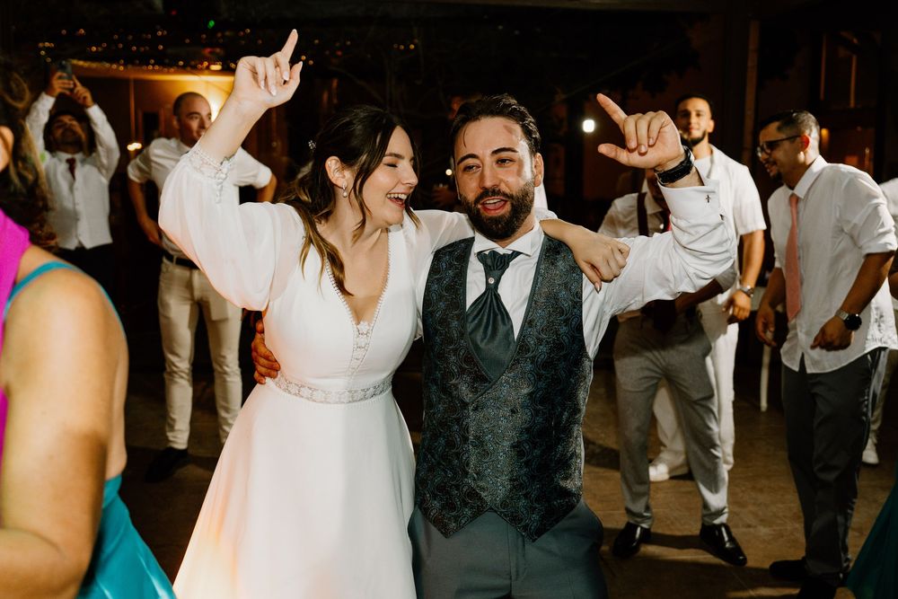 Joyful bride in a white gown and groom in a gray vest dance with raised arms as wedding guests celebrate behind them.