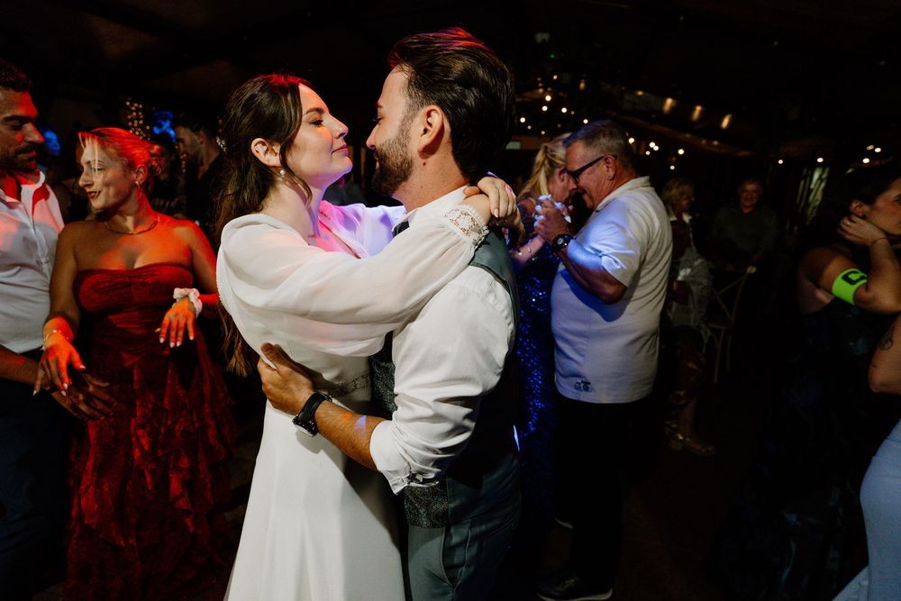 A bride and groom embrace and sway together on the dance floor while wedding guests celebrate around them under warm string lights.