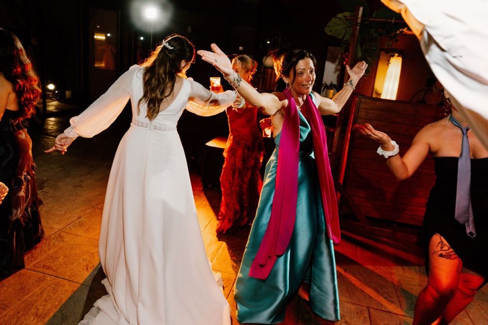 A bride in a white gown dances with a smiling guest in a teal dress and magenta scarf on the dance floor at a wedding reception.