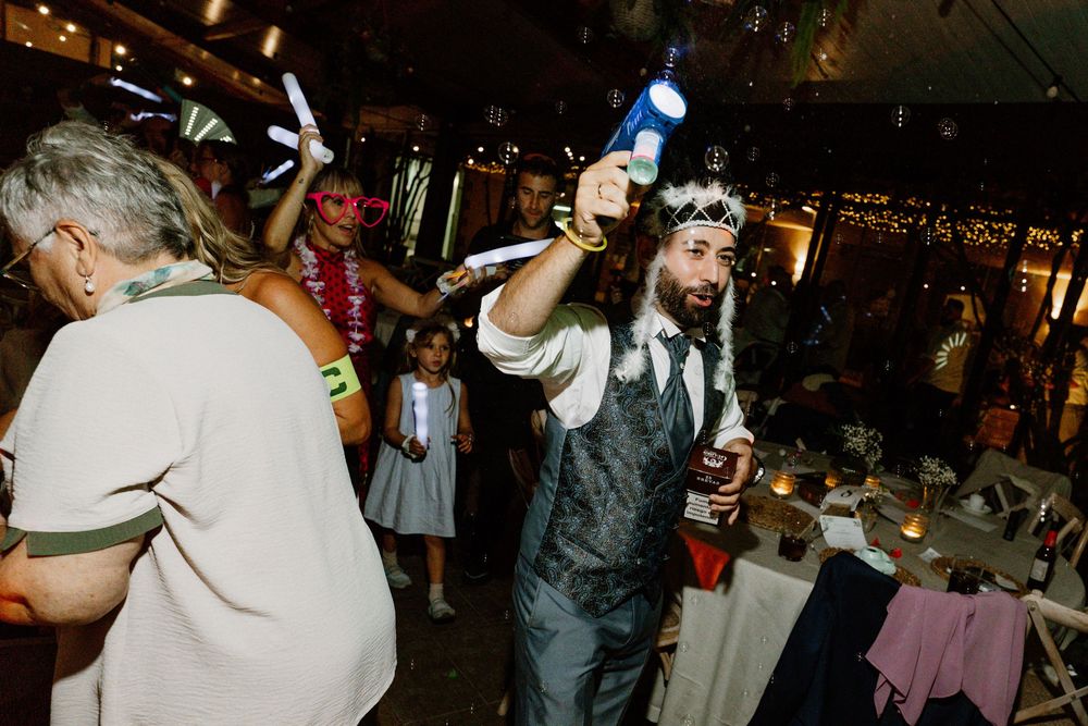 A bearded man in a vest and floral crown raises a glowing bottle while guests celebrate at a candlelit wedding reception.