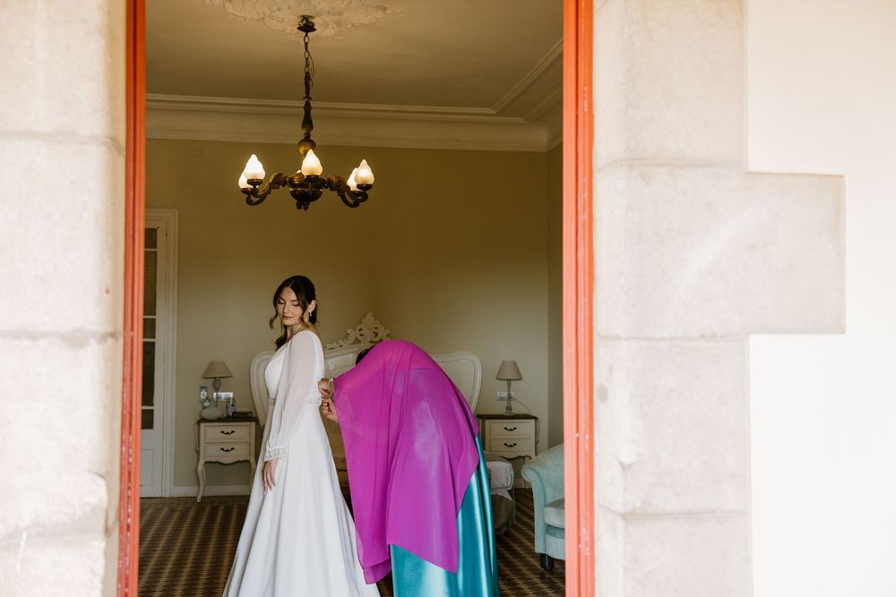 Bride in a white wedding dress stands in a bedroom doorway while a woman in a pink shawl helps her.