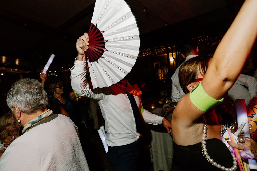 Guests dance at a lively evening party, one holding up a large white and red folding fan while a woman raises her arm in the foreground.