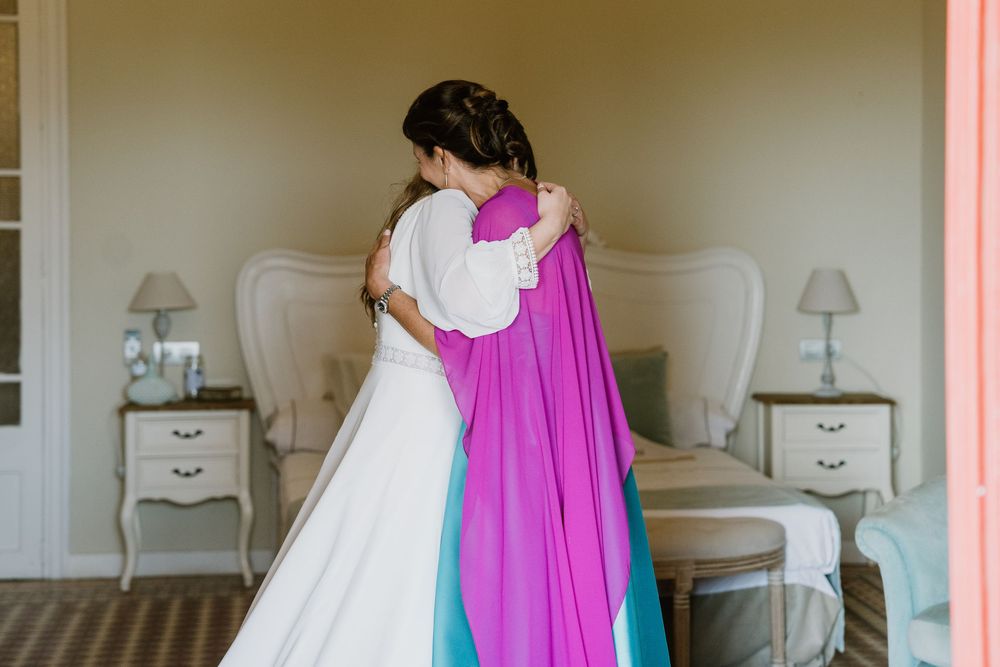 Bride in a white gown embracing a woman in a vibrant purple and teal dress in a bright, elegant bedroom with nightstands and lamps.