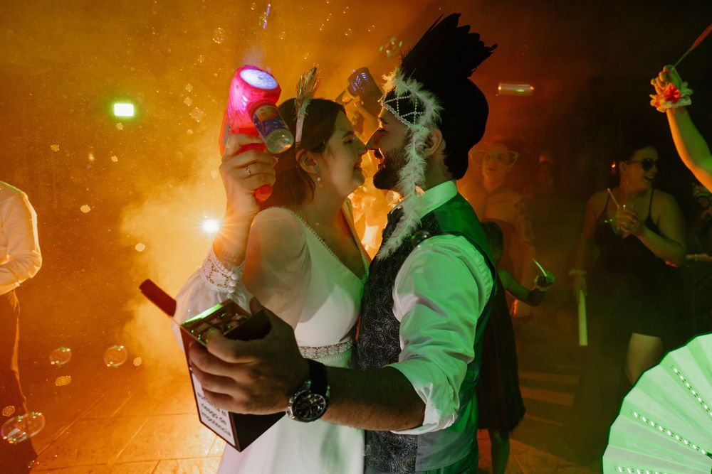 A bride in a white dress and a groom in a vest and crown kiss on a smoky dance floor while holding party props and a drink.