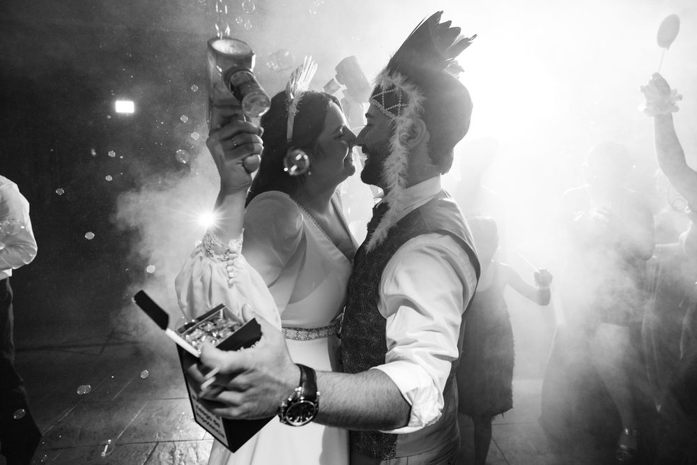 Black and white photo of a bride and groom kissing amid dancing guests and haze at a festive wedding reception party.