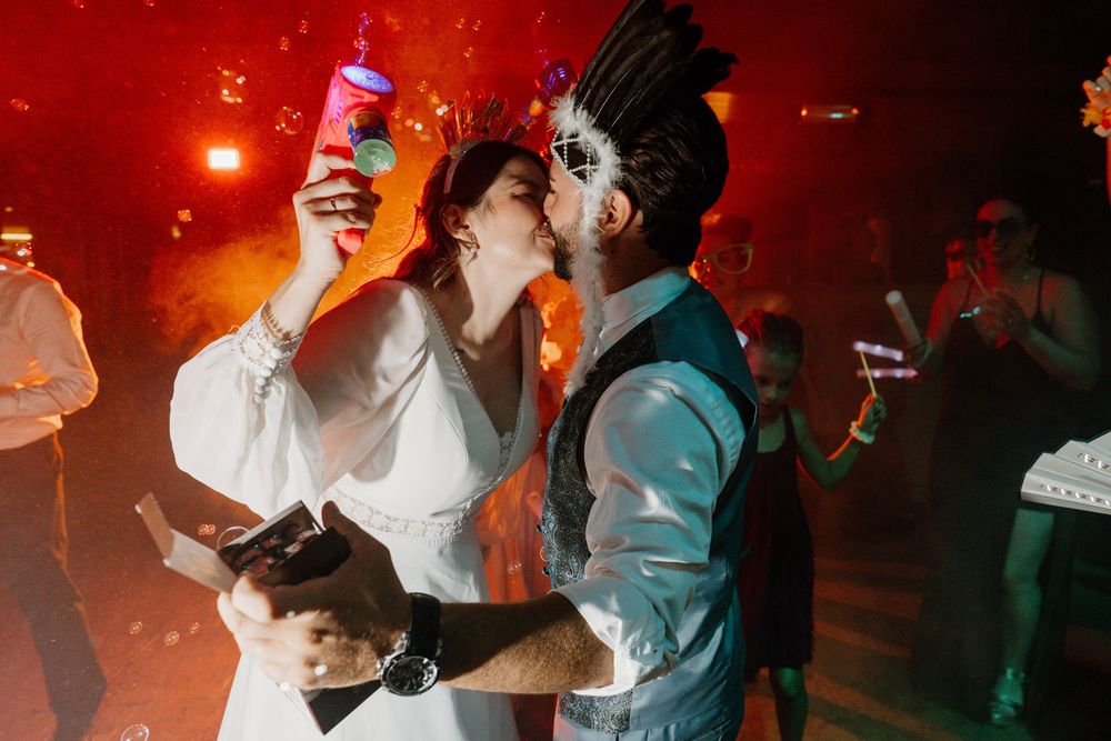 A bride in a white dress kisses a groom in a vest and sunglasses while guests dance and celebrate at a lively wedding reception.