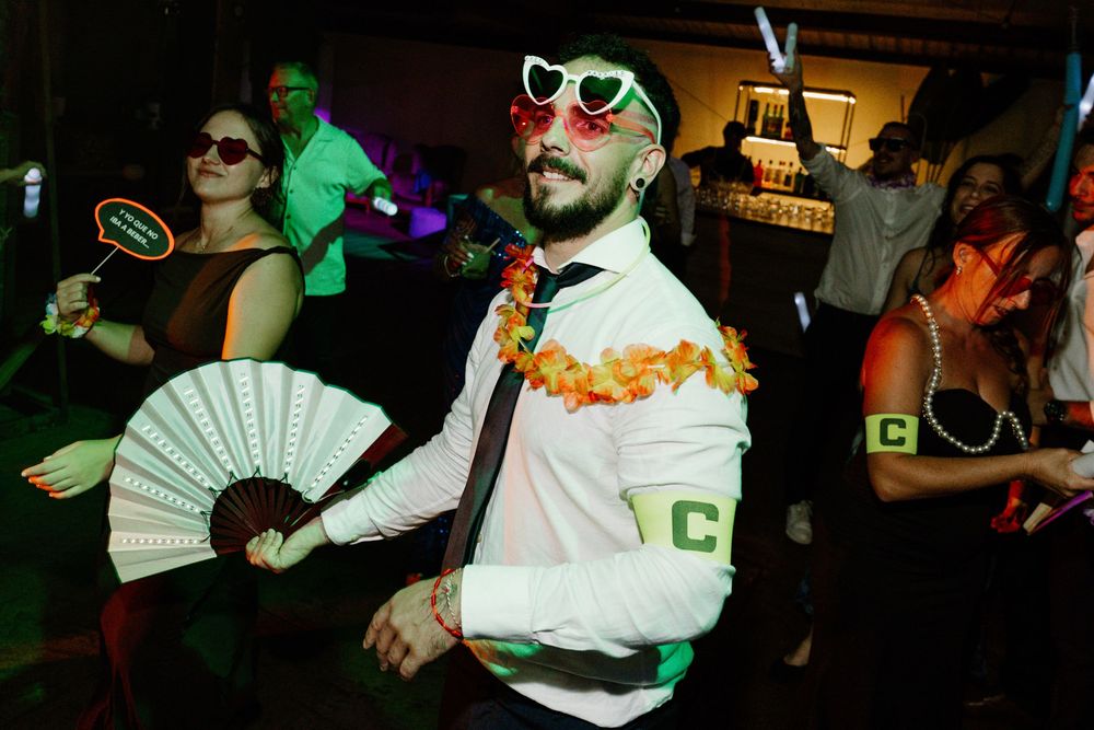 Guest in heart-shaped sunglasses and an orange floral lei dances at a wedding party while a woman nearby holds a white hand fan.