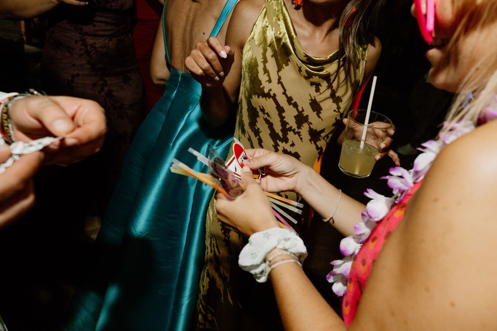 Guests at a lively party gather in a circle, one wearing a floral lei and wristband while holding and handing out small cards.