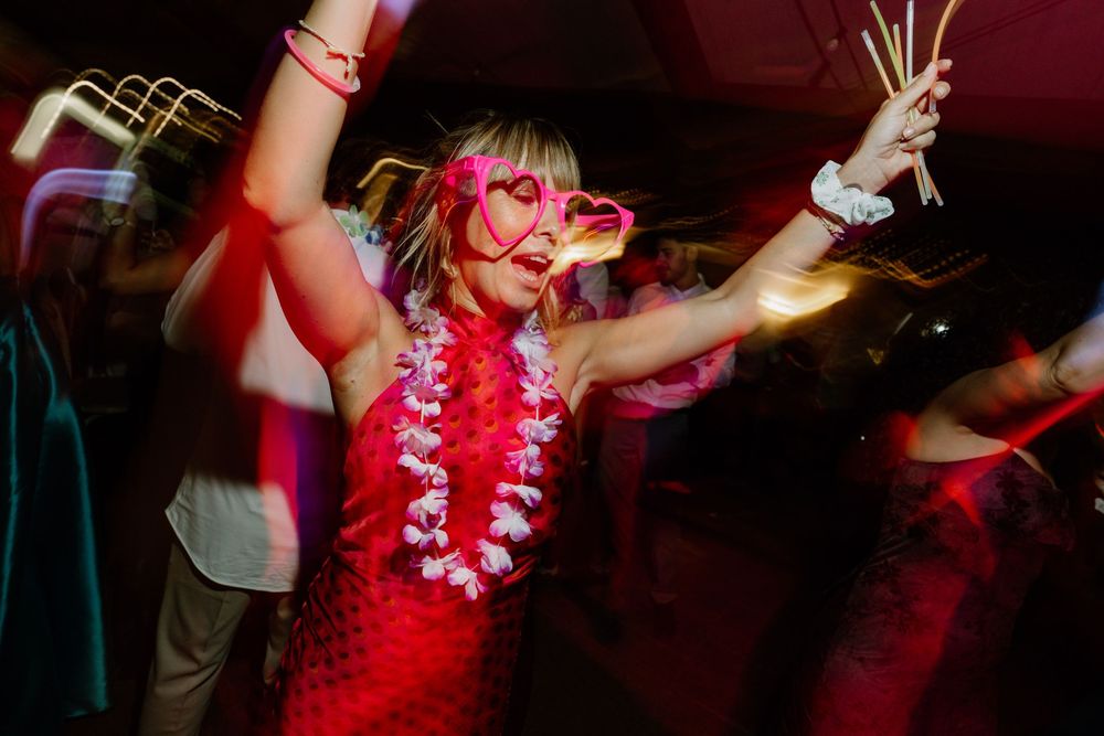Woman in a red dress and white flower lei dances joyfully at a lively party, wearing pink heart-shaped glasses with arms raised.
