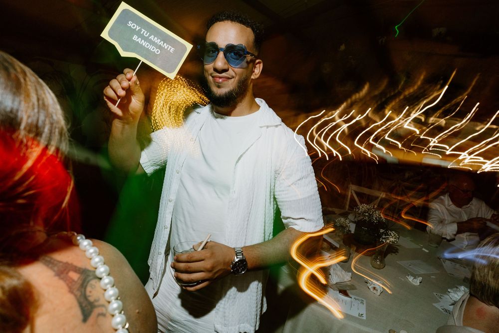 Smiling man in sunglasses and a white hooded jacket holds a handwritten sign while dancing at a lively evening party.