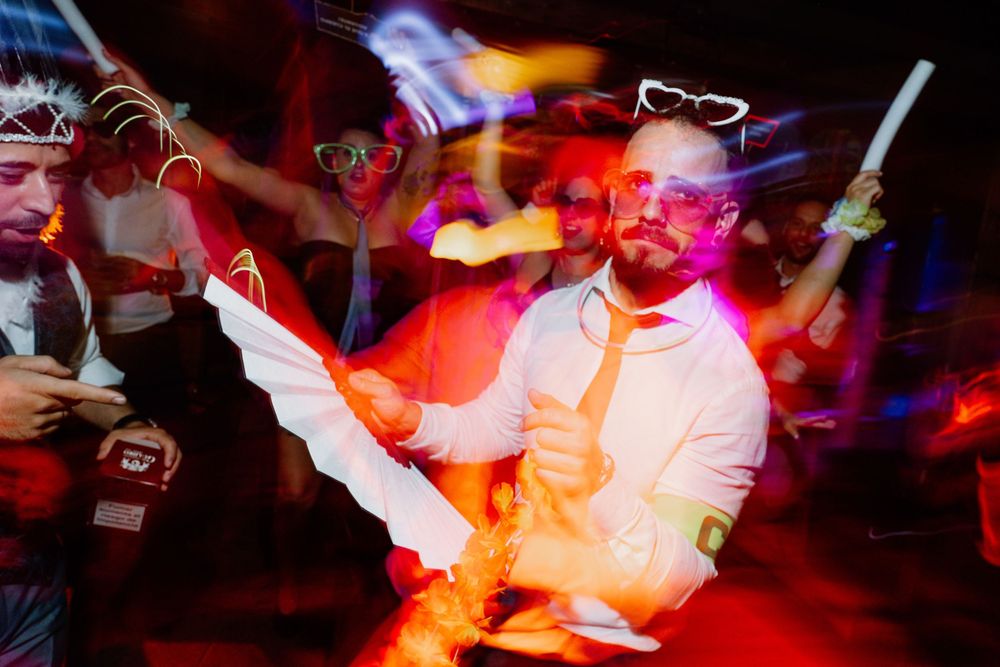 Guests in glow-in-the-dark glasses and neon accessories dance at a lively party, with motion blur and colorful lights filling the scene.
