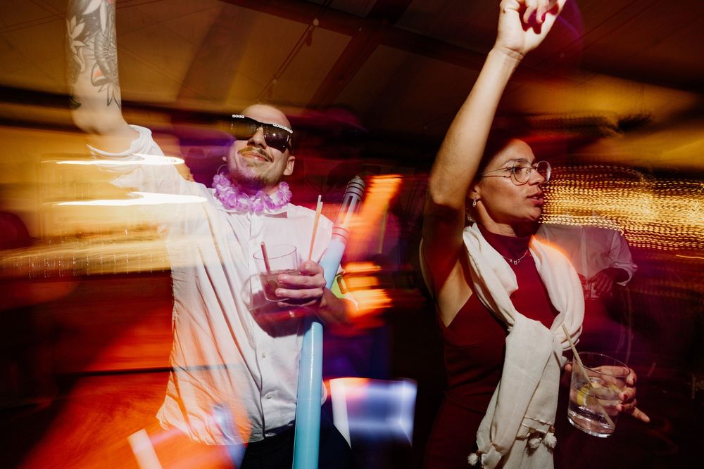 Two guests dance with raised arms at a dimly lit party, one wearing sunglasses and a pink lei, the other a red dress with white scarf.