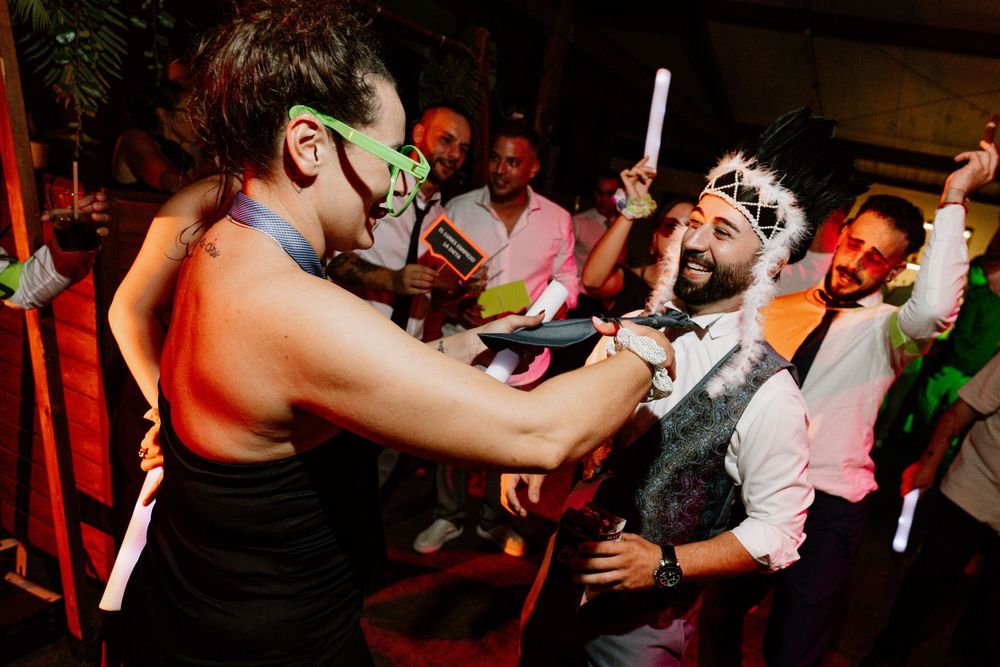 A guest in green glasses dances with a bearded man in a feather headdress as friends celebrate with glow sticks at a lively party.
