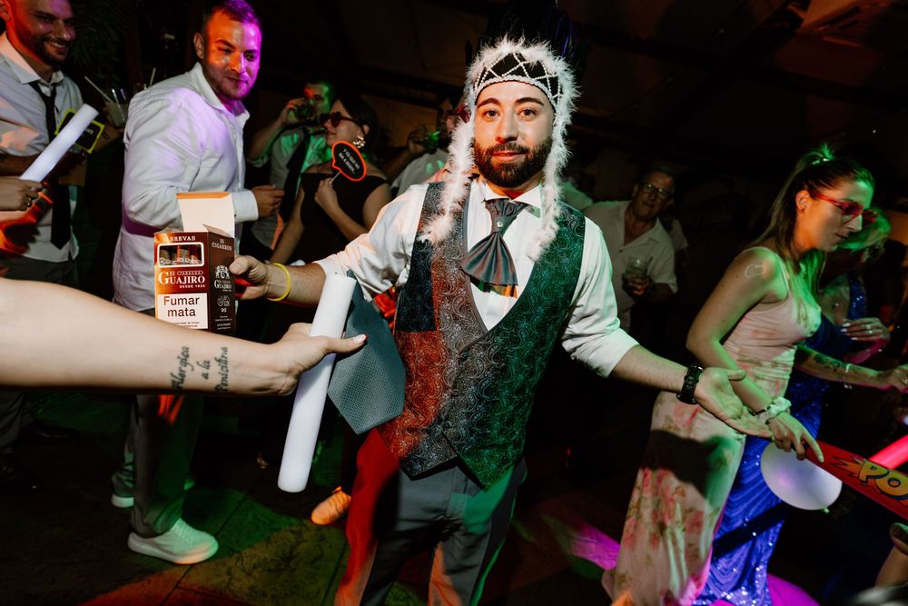 Bearded man in a feathered headdress, vest and tie dances energetically at a lively party while guests cheer and hold up drinks around him.