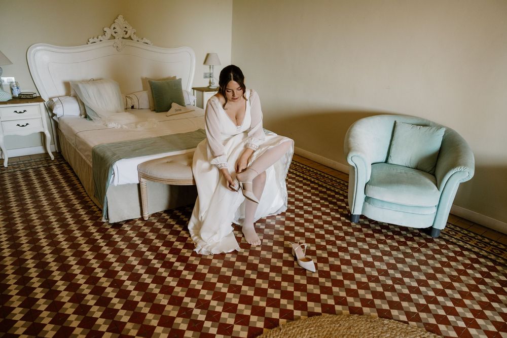 Woman in a white robe sits on a bench at the foot of a bed in an elegant hotel room with a checkered red and cream floor.