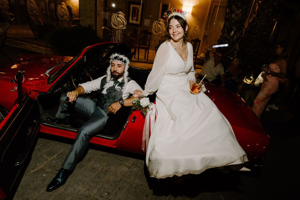Smiling bride in a white wedding dress stands beside a groom in a floral headpiece sitting in a red convertible car at night.