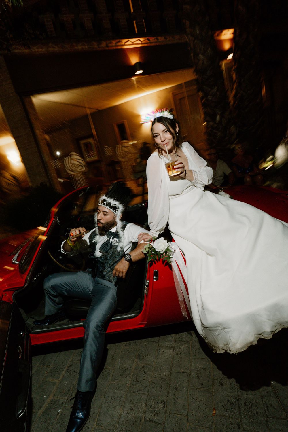 Bride in a flowing white gown and floral headpiece sits on a red convertible beside a groom in a vest and crown, sipping a drink at night.