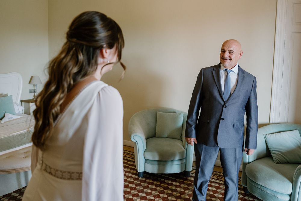 Bride in a white dress with a braided half-up hairstyle faces a smiling man in a gray suit in an elegant sitting room with teal armchairs.