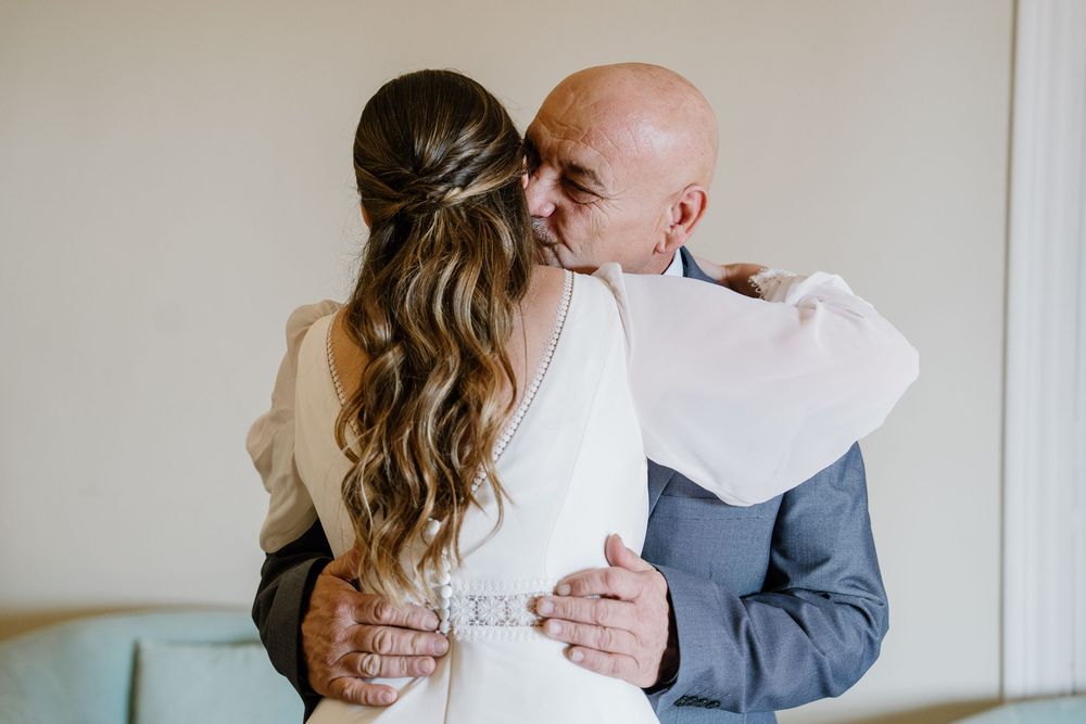 Bald man in a blue suit kisses the head of a bride in a white dress with a braided half-up hairstyle during an emotional wedding embrace.