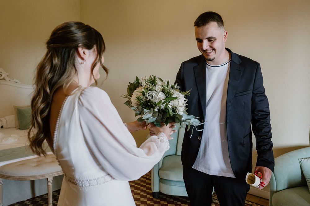 Smiling groom in a dark blazer looks at a bride in a white dress holding a bouquet of white flowers during a first look in a cozy room.