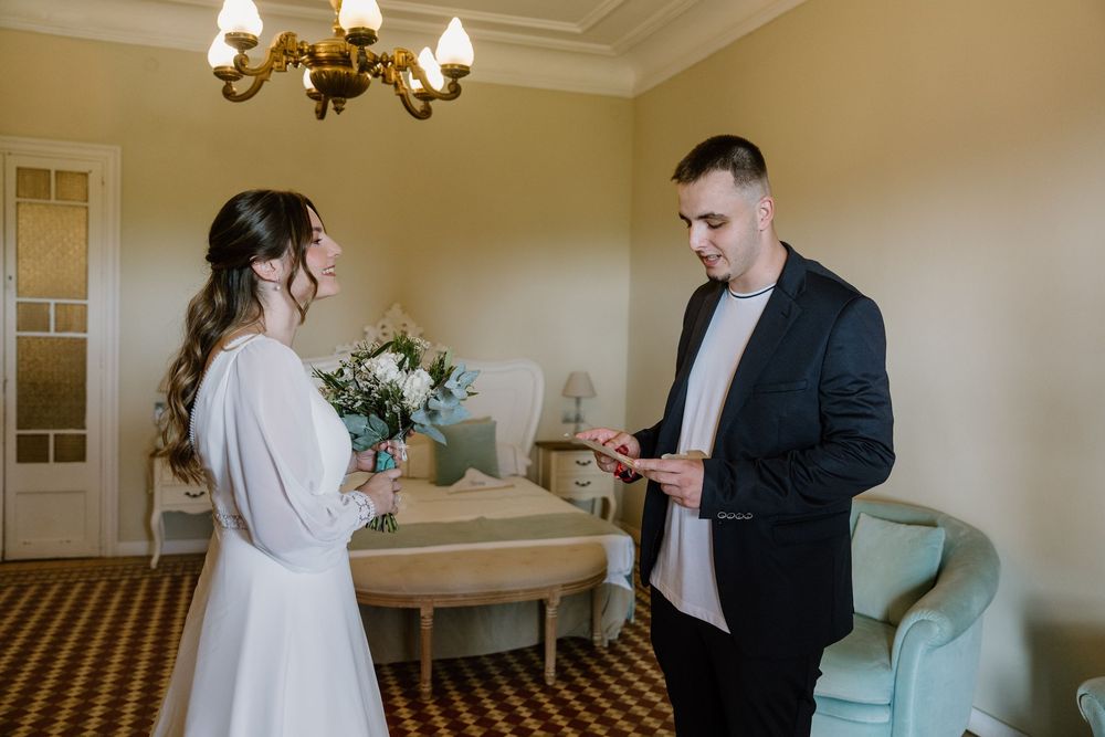Bride in a white dress holding a bouquet stands face to face with a groom in a dark blazer inside an elegant hotel room.