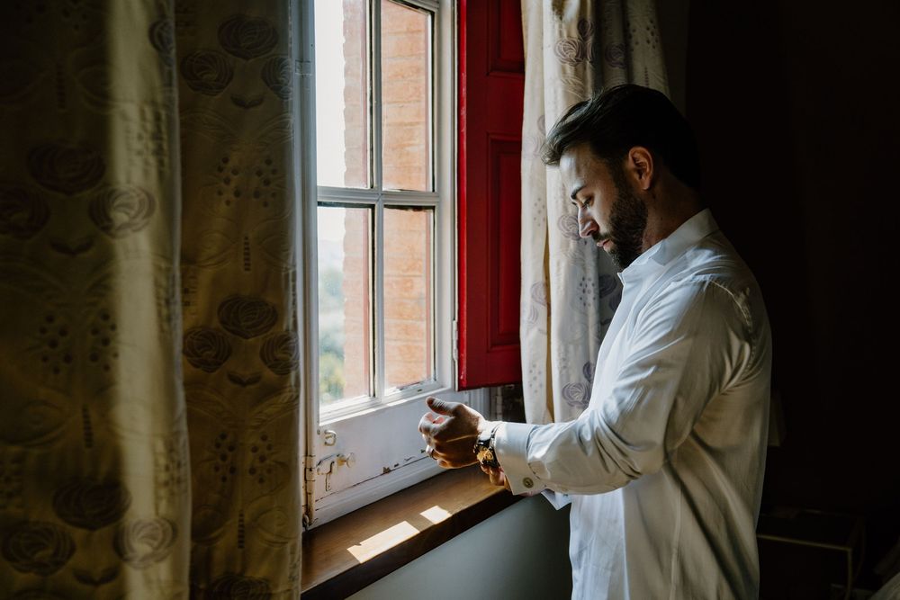 Bearded man in a white dress shirt stands by a sunlit window with patterned curtains, adjusting his cuff and watch.