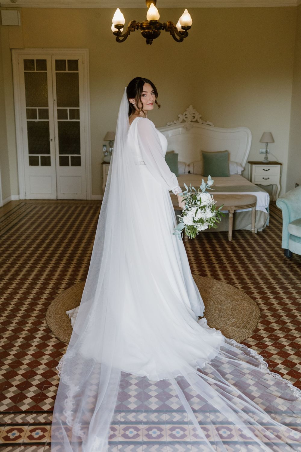 Bride in an elegant white gown with a long flowing veil holds a white floral bouquet in a bright vintage hotel suite.