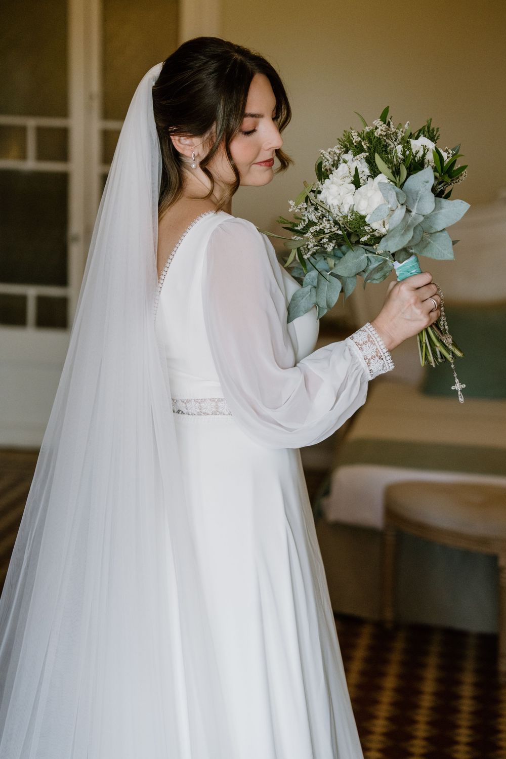 Smiling bride in a white long-sleeve gown and flowing veil holds a green and white bridal bouquet in a classic room.