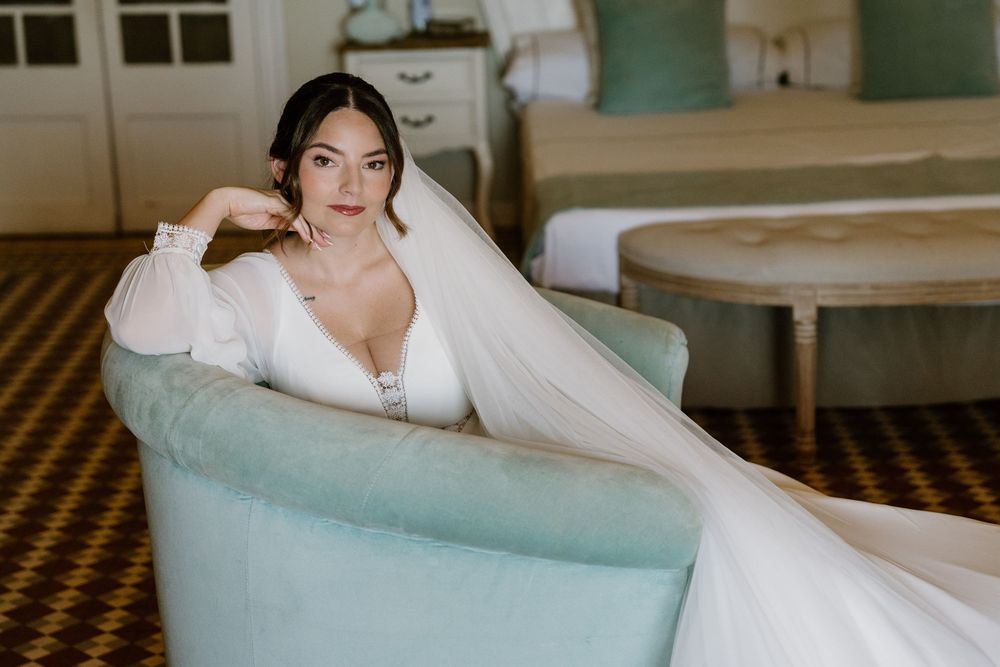 Bride in a white long-sleeve gown and veil poses seated in a mint green armchair in an elegant bridal suite.