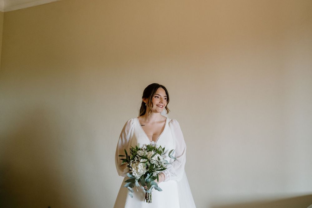 Bride in a white wedding dress holding a green and white bouquet while gazing to the side against a soft neutral wall.