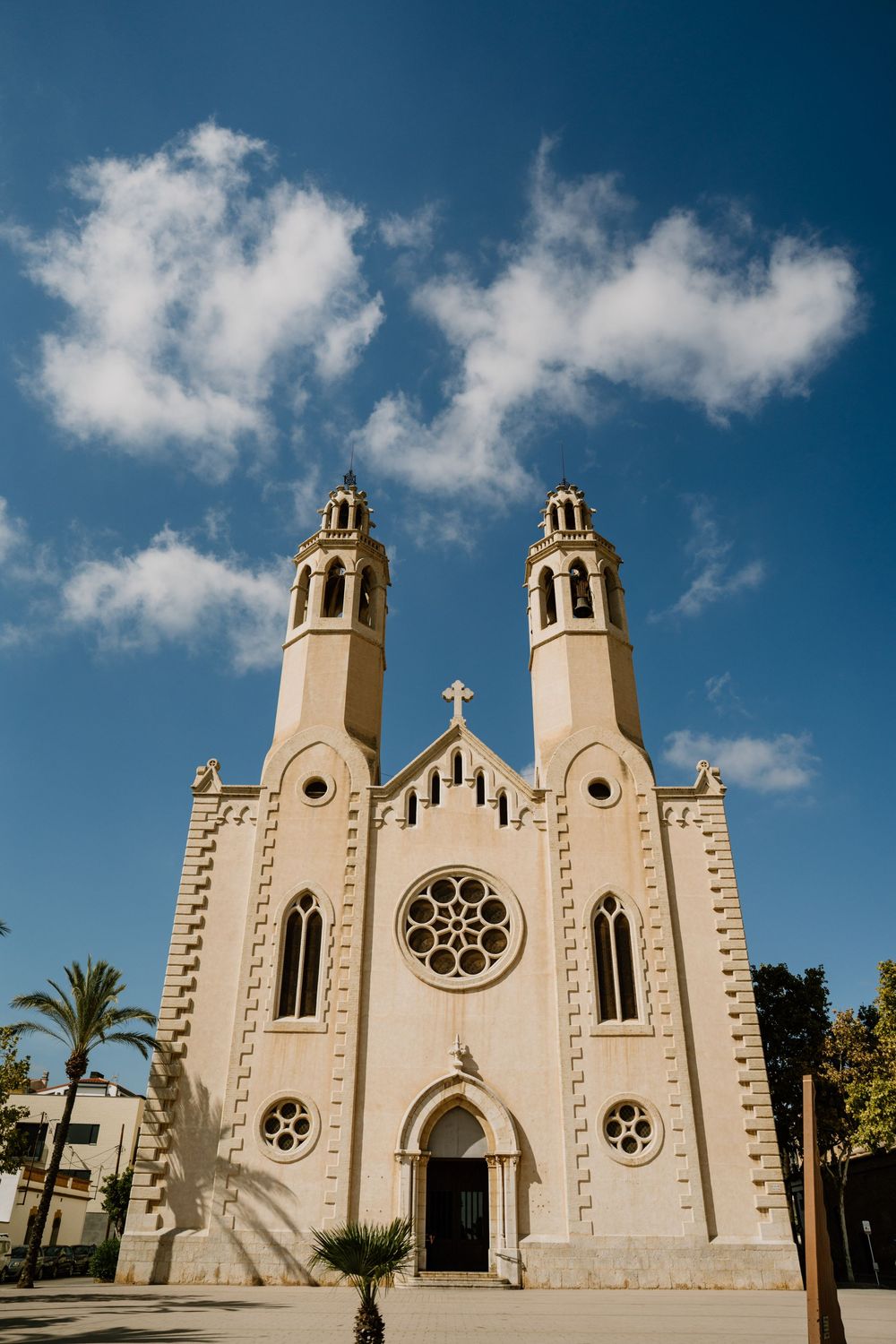 Cream-colored neo-Gothic church with twin bell towers, a rose window and arched entrance under a blue sky with palm trees.
