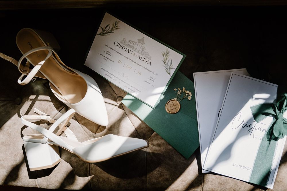 White pointed-toe bridal heels beside a green and white wedding invitation suite with a gold wax seal, lit by soft sunlight.